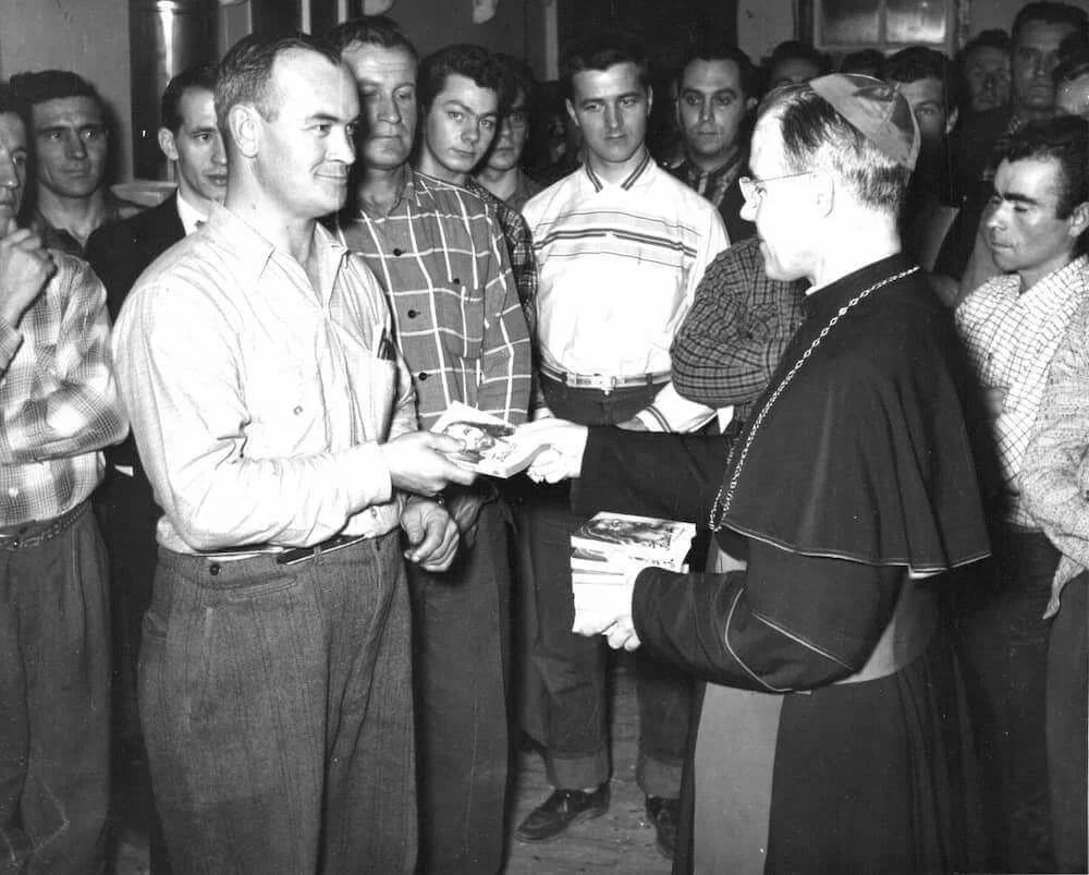 Mons. Couturier entrega un documento a un trabajador, rodeado de leñadores